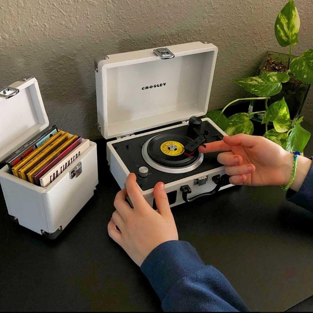 The image shows hands adjusting the tonearm of the Crosley Mini Cruiser Turntable - White Sand, a compact and portable vinyl player, next to a matching vinyl case