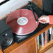 The image shows the Crosley T150C Shelf System - Black playing a pink vinyl record, with a person adjusting the tonearm near a wooden shelf filled with records