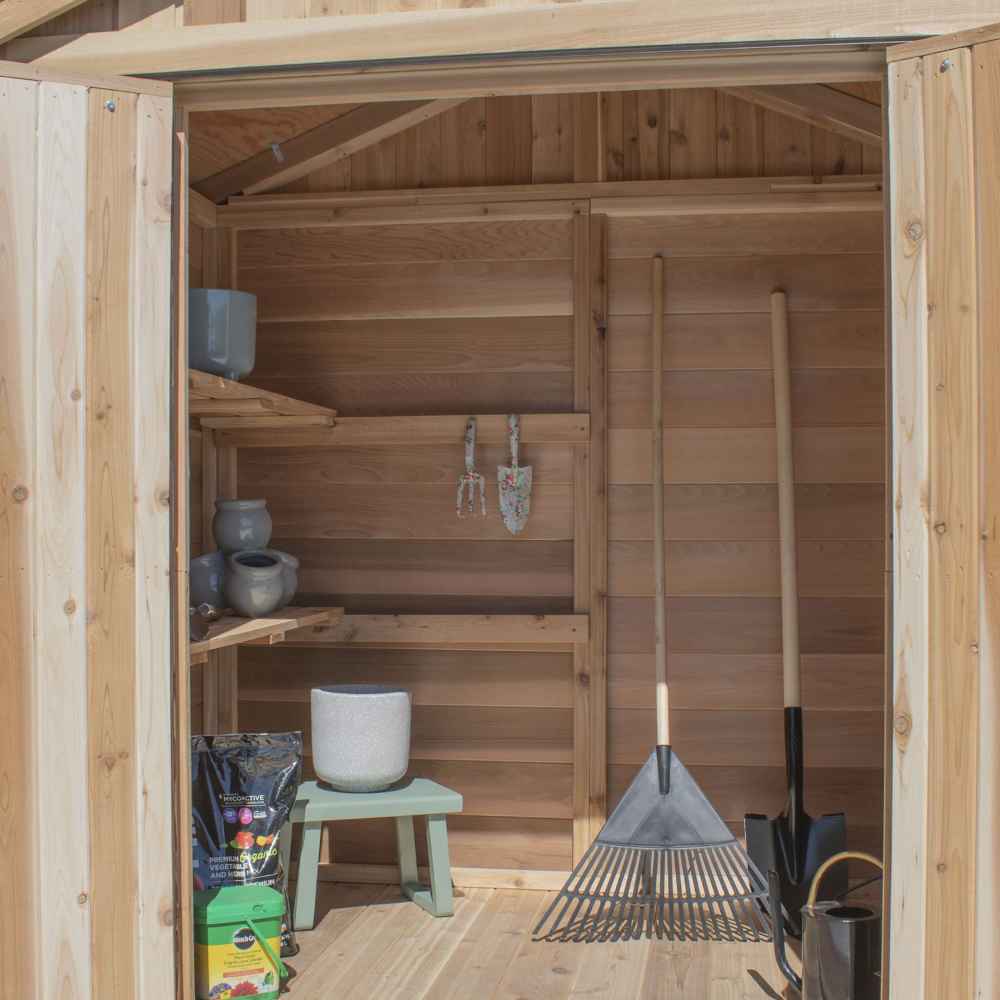 The interior of the Outdoor Living Today SpaceMaster 6×4 with Double Door shows shelves, garden tools, pots, and rakes neatly stored inside the wooden shed