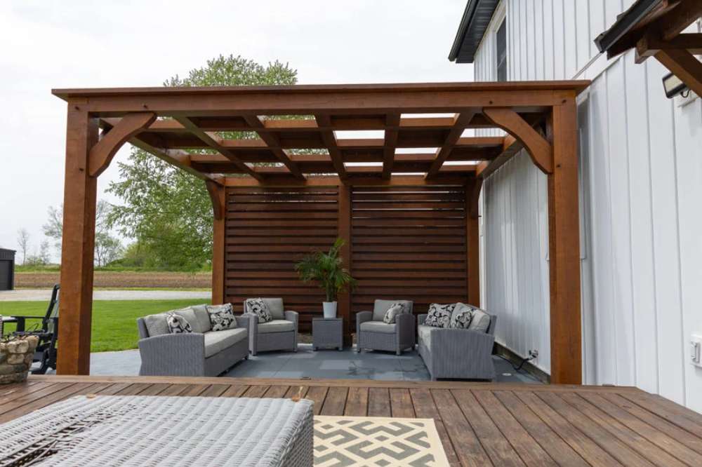 This Brookside Timber Frame Timber Resort Pergola offers a cozy and stylish outdoor retreat with a slatted privacy wall and elegant timber craftsmanship