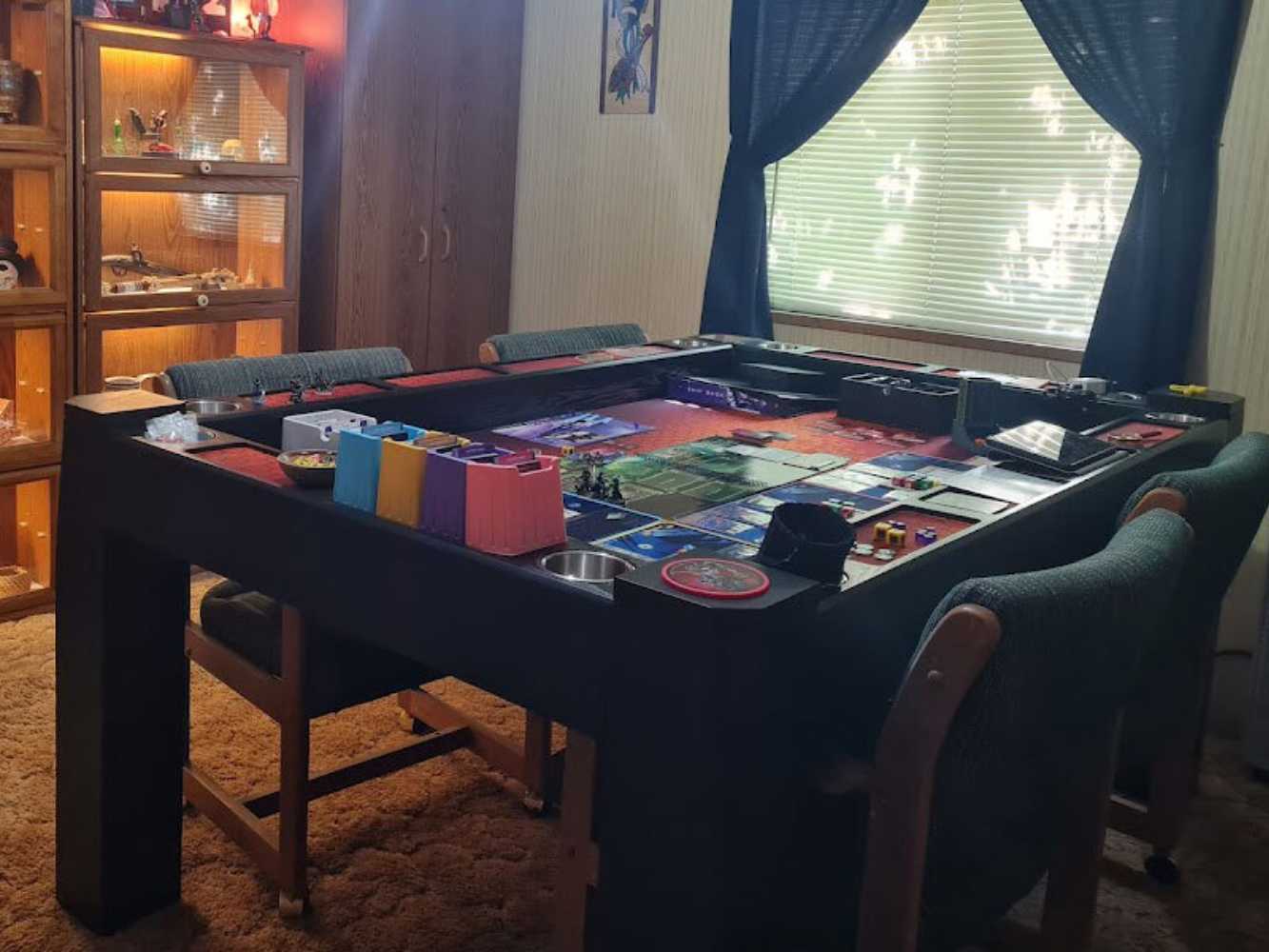 This Game Theory Tables Origins Lit Game Table is surrounded by cozy chairs and set up with colorful game components