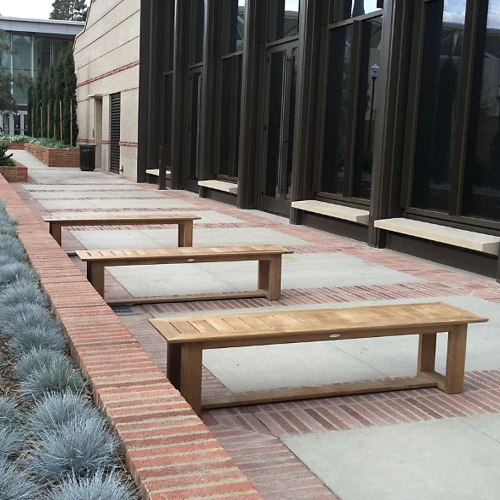 Three evenly spaced wooden benches, each a Westminster Teak 6ft Horizon Teak Backless Bench, are lined along a brick-edged walkway beside a modern building with tall windows