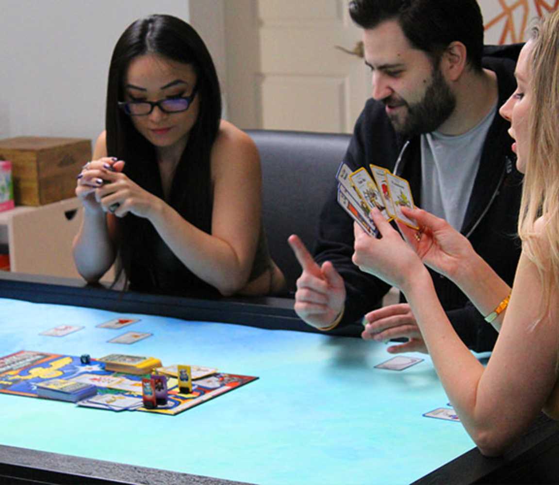 Three friends enjoy a board game on the Game Theory Tables The Meeples Champion Game Table, with a bright playing area