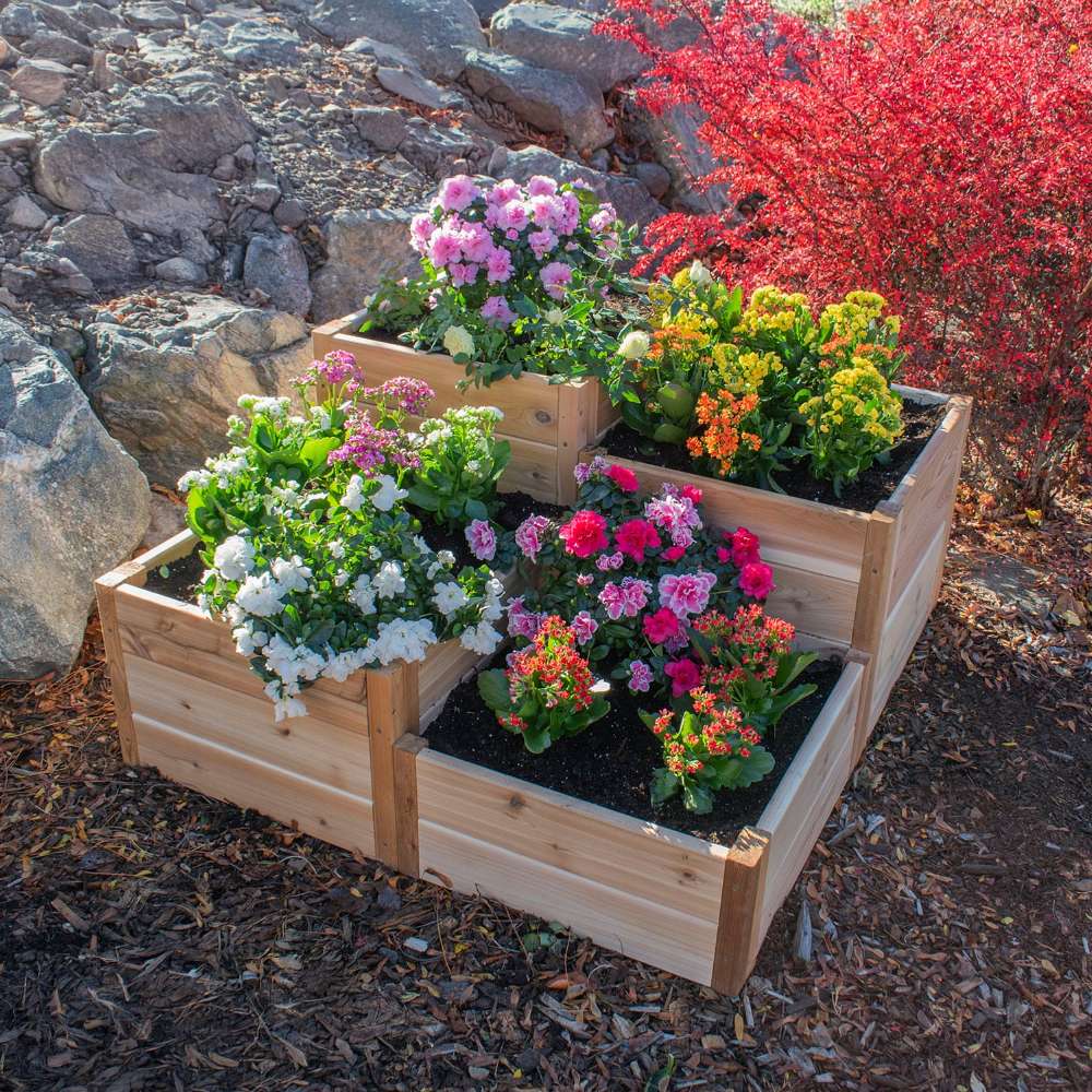 Tiered cedar planter filled with colorful flowers sits among rocks, featuring the Outdoor Living Today 3.5×4 Tiered Cedar Corner Raised Garden Bed at the center