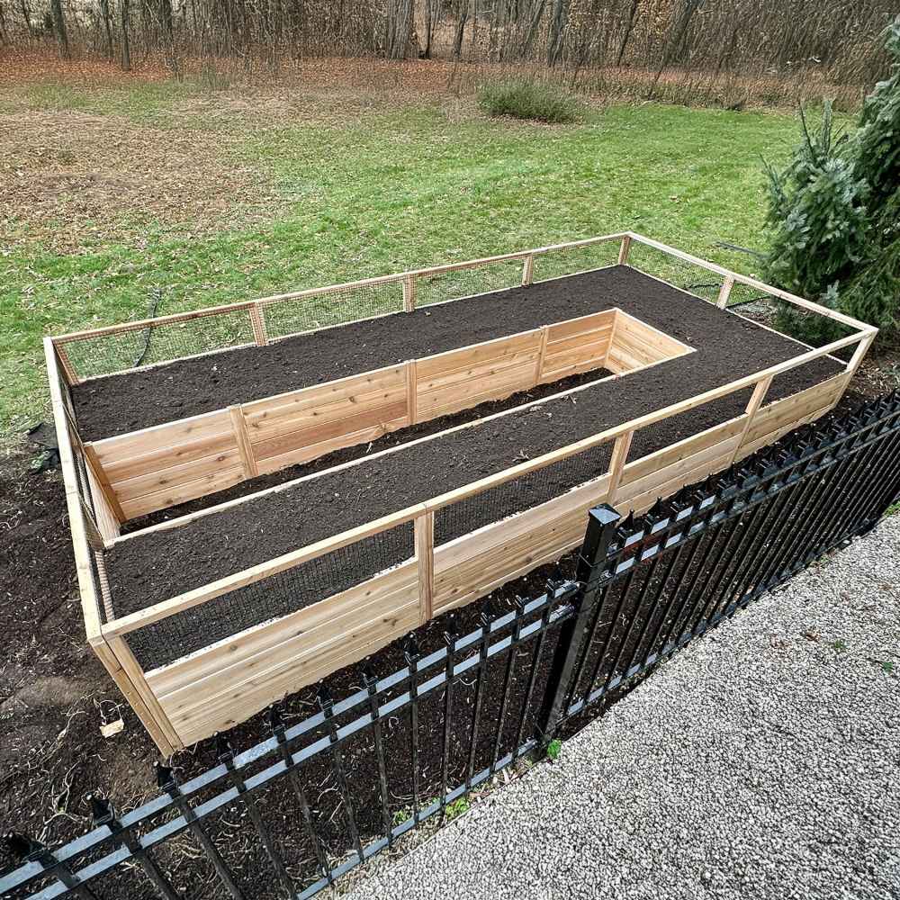 Top view shows the Outdoor Living Today 8×16 Raised Garden Bed filled with soil, framed by a black metal fence and surrounded by grass