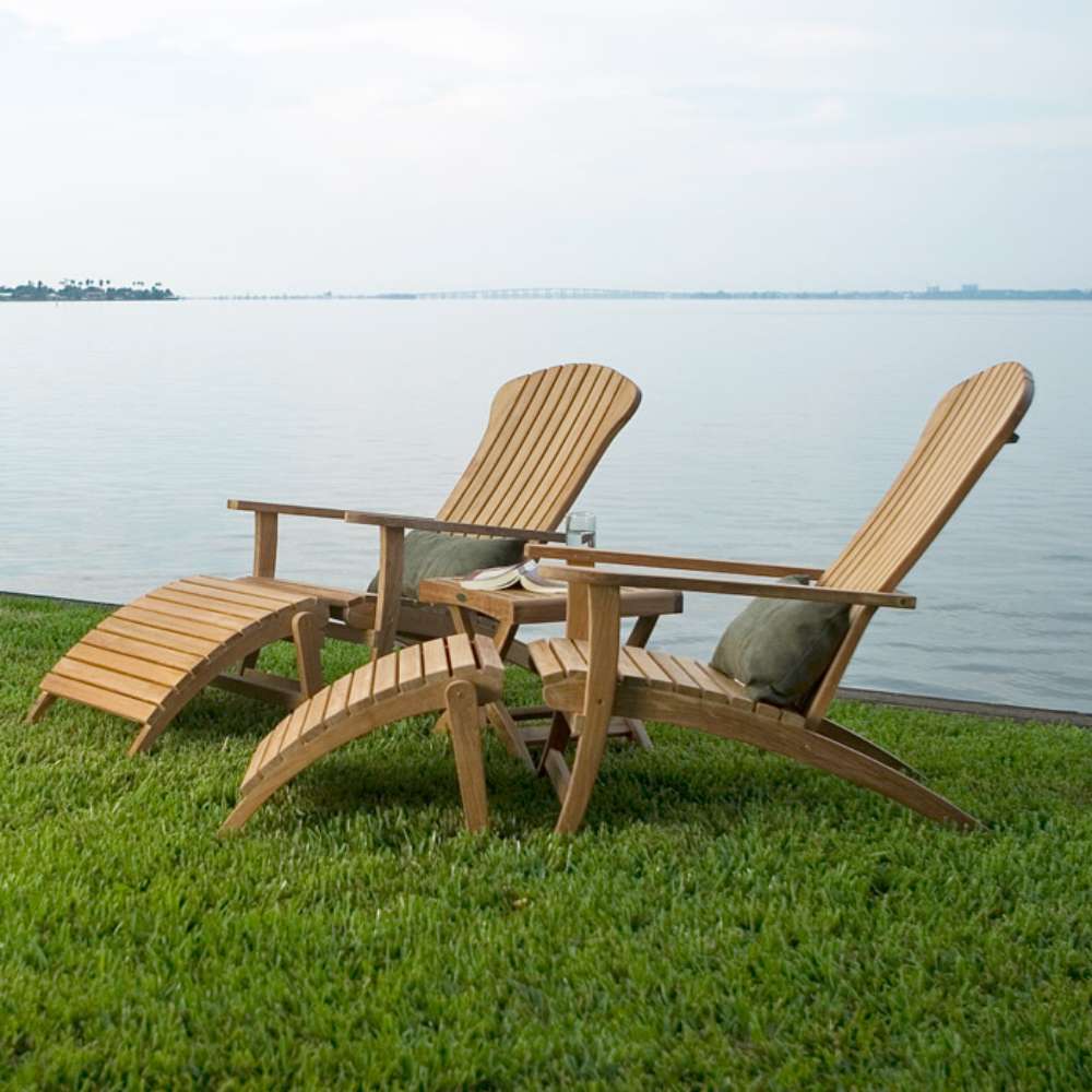 Two Westminster Teak Adirondack Lounger and Ottoman chairs with cushions sit on green grass beside calm water for relaxing outdoor seating