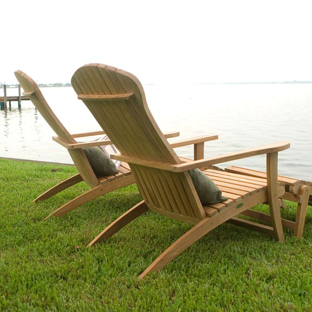 Two Westminster Teak Adirondack Lounger and Ottoman chairs with matching footrests are placed on grass beside calm water for outdoor lounging