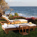 Two Westminster Teak Horizon Chaise Lounger chairs with cushions sit on a grassy area by the ocean, accompanied by small wooden side tables and a sun hat