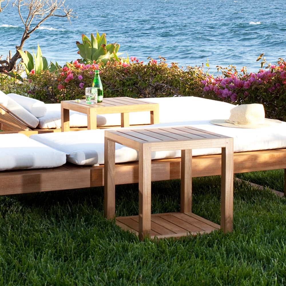 Two Westminster Teak Horizon Teak Side Tables sit on grass beside cushioned loungers with an ocean view in the background