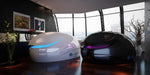 Two Dreampod V2 Float Pods, one white with a blue accent and one black with a purple accent, sit side-by-side in a modern, high-rise room with large windows