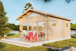 Two bright red chairs sit on the front porch of a cozy wood cabin beside a lake, showcasing the Sawmill Structures Edwin Lake House 70MM Garden Room