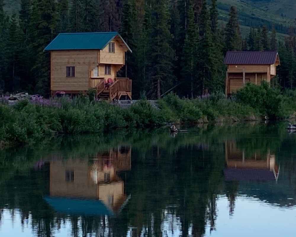Two wooden cabins reflect off a still lake at dusk, with the left cabin being the Sawmill Structures The Jumbo Grand Loft 130 SD Garden Room