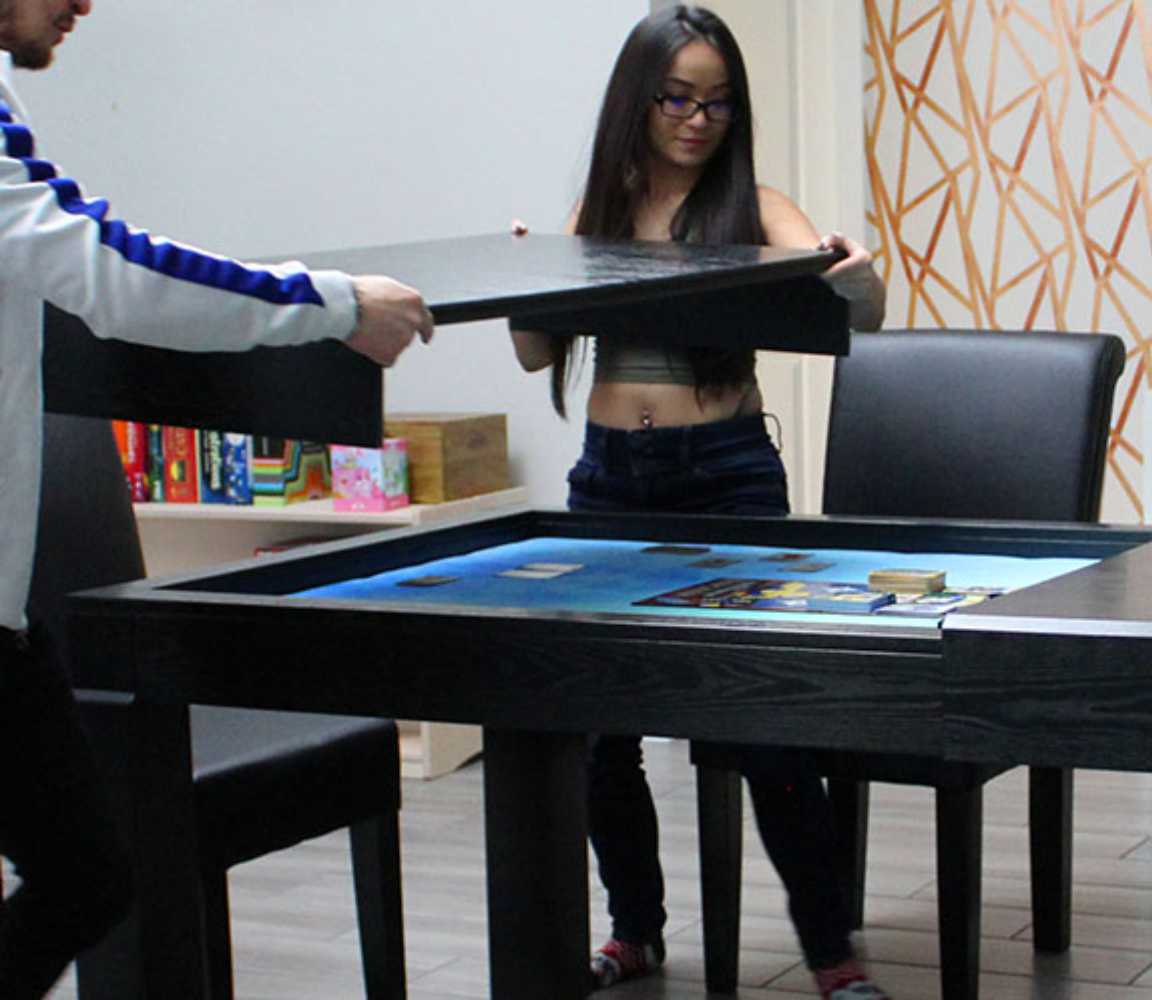 Two people assemble the Game Theory Tables The Meeples Champion Game Table by lifting its dining top, revealing a beautiful blue playing area underneath