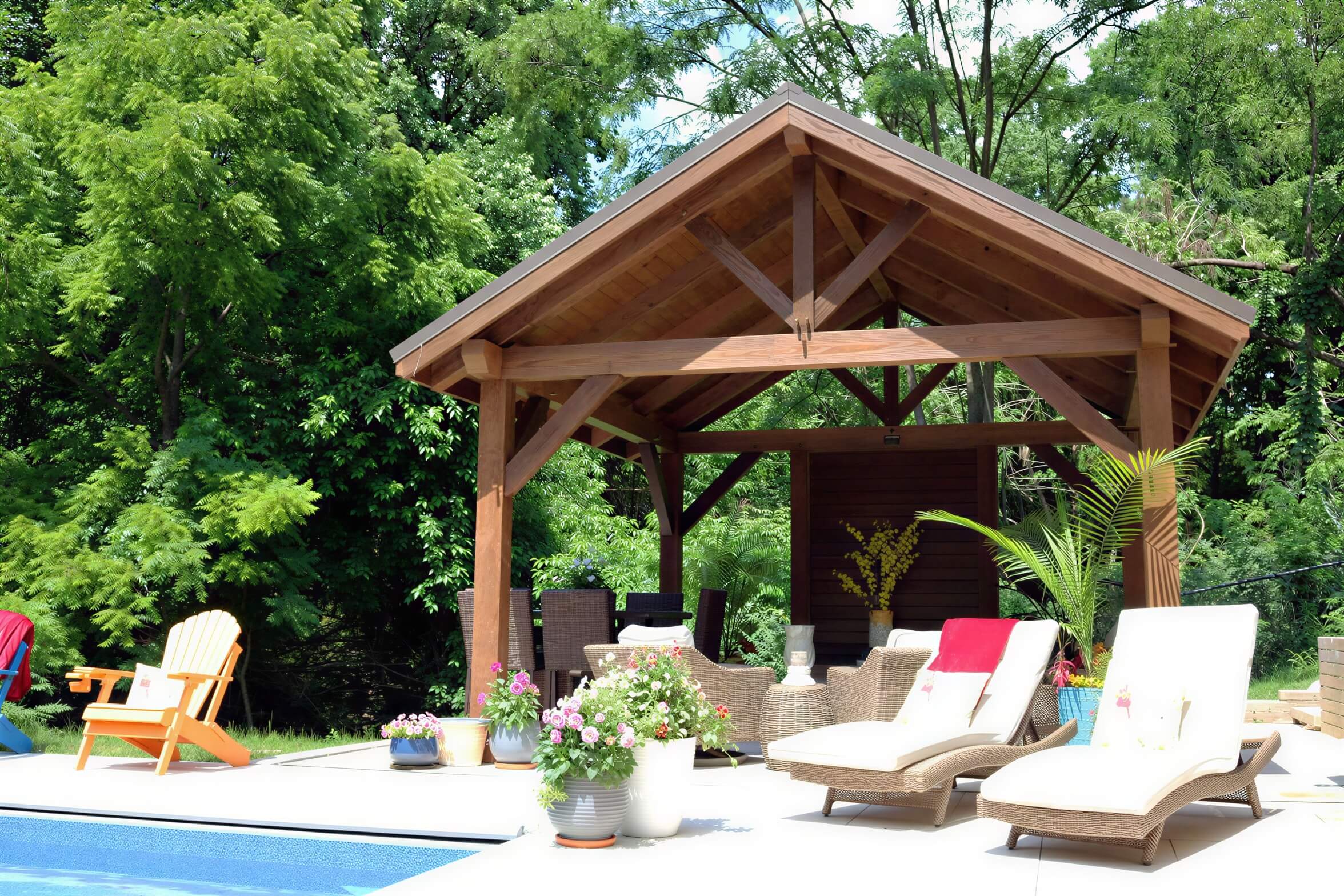Under Brookside Timber Frame Forest Retreat Pavilion, comfortable lounge chairs and plants create a peaceful poolside retreat surrounded by dense greenery
