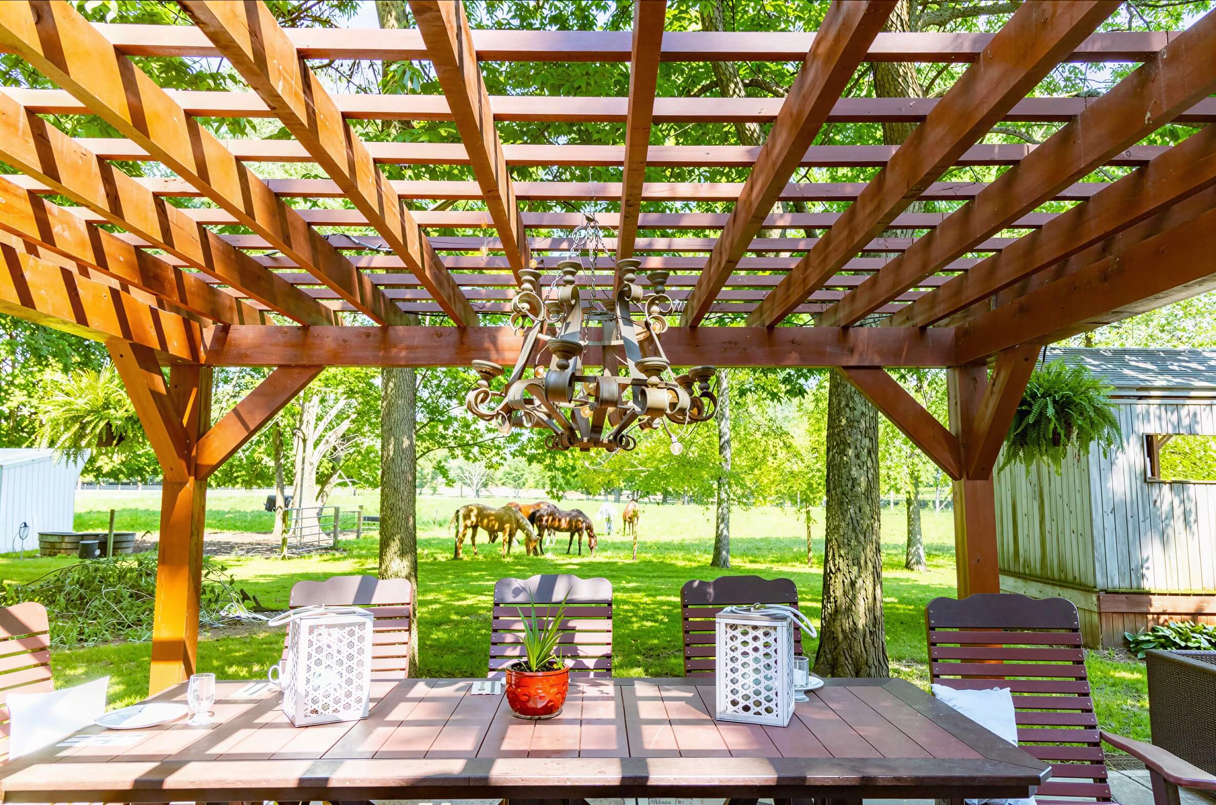 Under the Brookside Timber Frame Fern Shade Pergola, a dining table sits beneath a decorative chandelier, with sunlight filtering through the wooden slats above