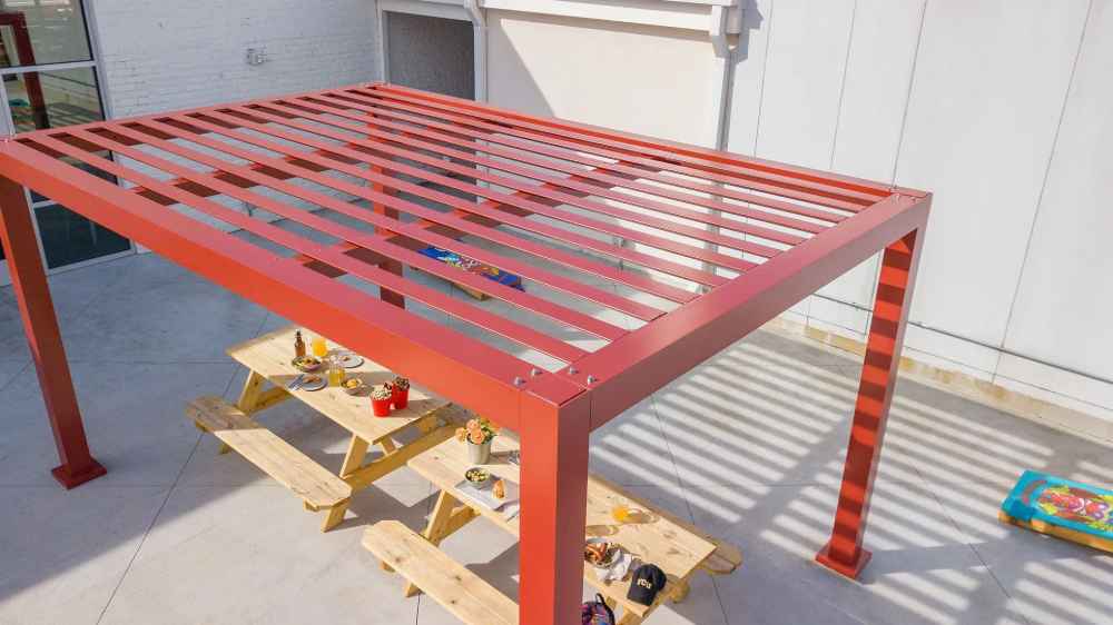 Under the open slats of a red Structureworks Element Trex Pergola, several wooden picnic tables are arranged for a sunny outdoor gathering