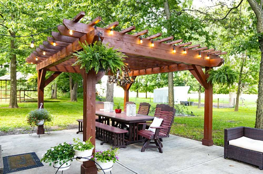 Under the Brookside Timber Frame Shadelan Retreat Pergola, a large wooden dining table with cushioned chairs invites guests to relax, surrounded by lush greenery and rustic decor