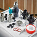 Valves, hose connectors, a drain grate, red O-rings and white thread seal tape for Golden Designs Dynamic Cold Therapy Cuboid Plastic Cold Plunge Tub are spread across a white surface