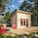 Angled view of the Versa 107 70MM on a spacious wooden deck with glazed doors, a side window, exterior lights, red chairs, and surrounding pine trees.