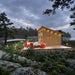 Versa 240 70MM garden room at dusk beside a lake, illuminated by warm exterior and pathway lights with red Adirondack chairs on the wooden deck.