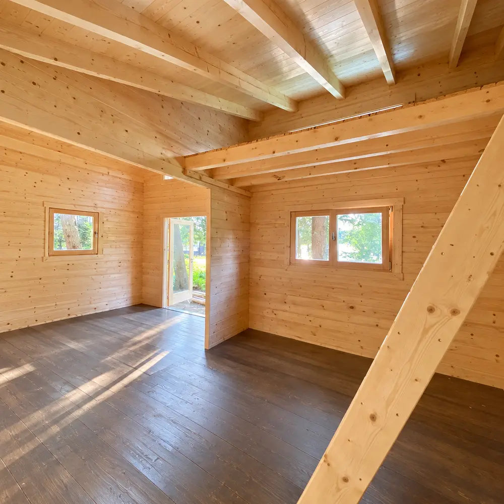 Unfinished Versa 240 70MM interior with exposed wooden roof beams, dark flooring, multiple windows, an open doorway, and a diagonal timber ladder.