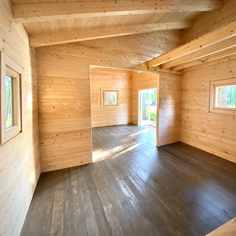 Unfinished Versa 240 70MM interior showing two connected rooms with natural wood walls, dark flooring, exposed ceiling beams, windows, and a bright exterior doorway.