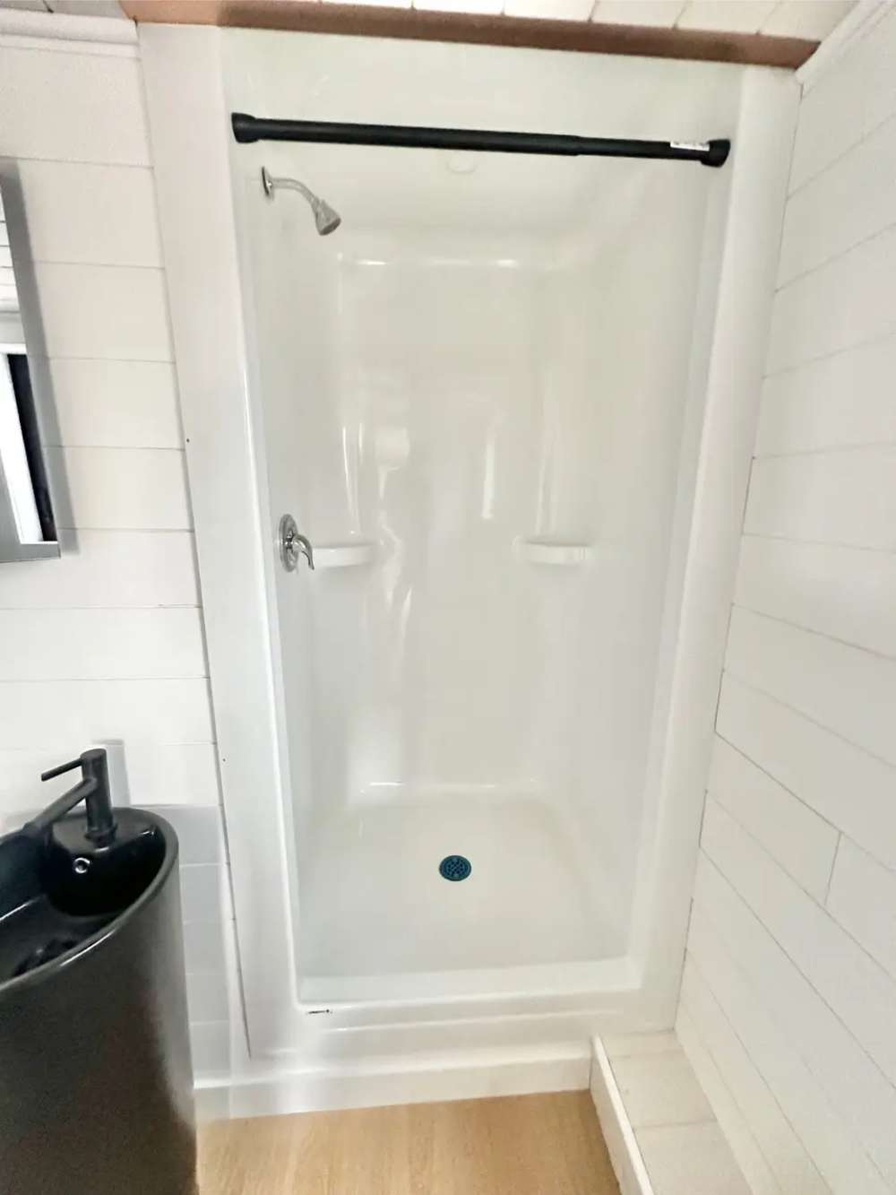 Walk-in shower in Sawmill Structures Wanderlove Tiny Home is white, compact, and framed by light shiplap walls with a black curtain rod
