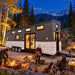 Wanderlove House on Wheels glowing at a Colorado forest campsite after sunset, as three adults prepare warm drinks and relax beside a contained campfire with snow-dusted mountains behind them.