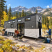 Wanderlove House on Wheels at a sunny Colorado mountain campsite, where three adults unpack hiking equipment, arrange camp chairs, and review a trail map beneath the Rocky Mountains.