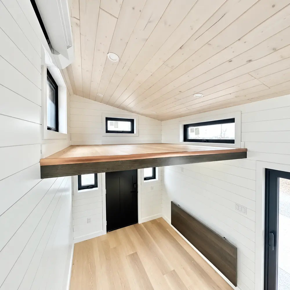 Loft above the Wanderlove Tiny Home entrance with white shiplap walls, light wood flooring, several black-framed windows, and a low ceiling.