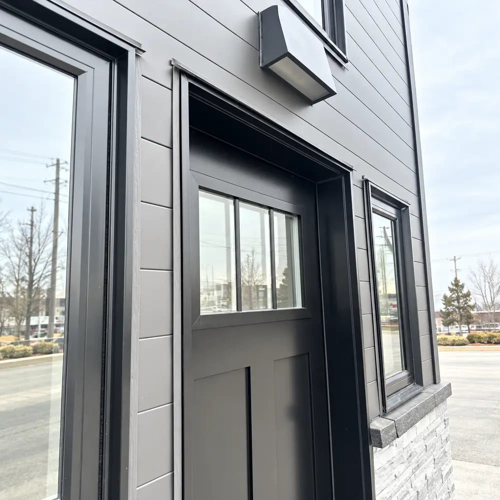 Exterior close-up of the Wanderlove Tiny Home’s black front door framed by dark siding, narrow windows, and a rectangular wall light.