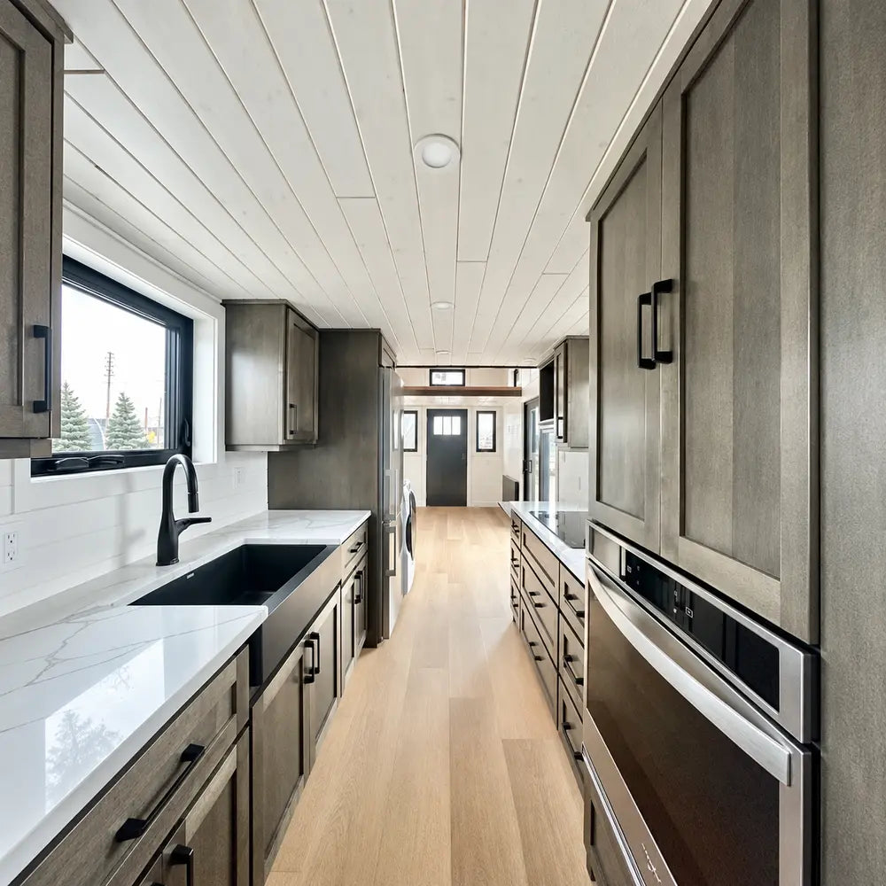 Long galley-style kitchen inside the Wanderlove Tiny Home with dark cabinetry, marble-style counters, appliances, windows, and light wood flooring.