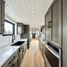 Long galley-style kitchen inside the Wanderlove Tiny Home with dark cabinetry, marble-style counters, appliances, windows, and light wood flooring.