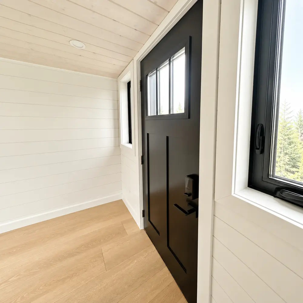 Interior view of the Wanderlove Tiny Home’s modern black front door with vertical panels and glass panes beside white shiplap walls.