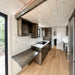 Wanderlove Tiny Home kitchen with dark wood cabinets, black cooktop, marble-style counter, small work desk, and exposed wood ceiling beam.