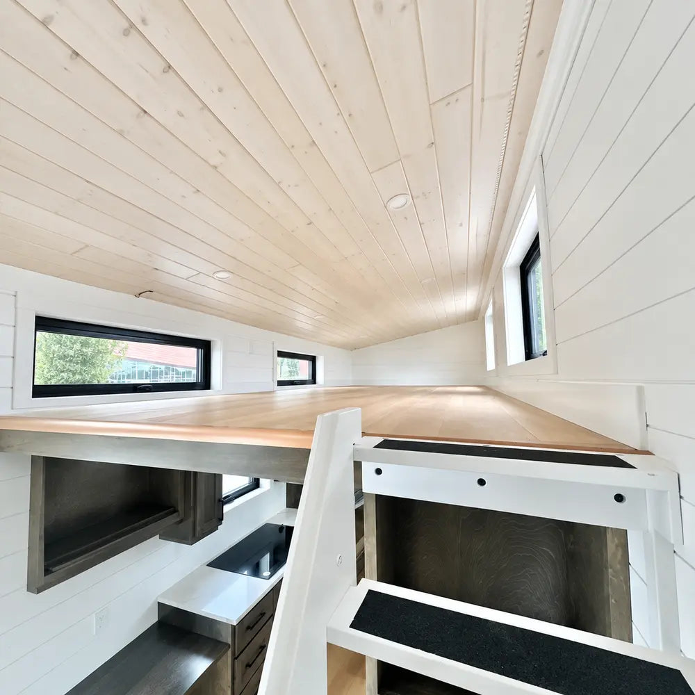 Low-ceiling loft inside the Wanderlove Tiny Home with white shiplap walls, light wood surfaces, several windows, and compact staircase access.