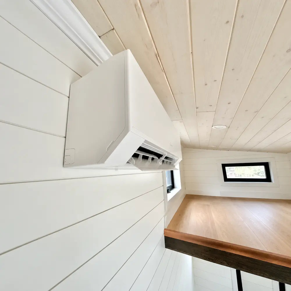 White mini-split air conditioner mounted high on a shiplap wall beside black-trimmed windows inside the Wanderlove Tiny Home.