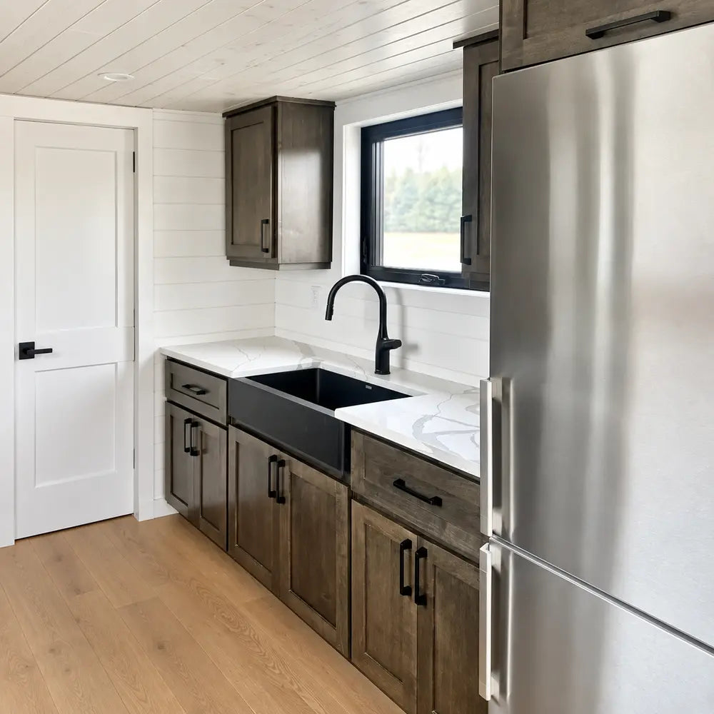 Wanderlove Tiny Home kitchen with a black sink, marble-style countertop, stainless steel refrigerator, dark cabinetry, and window.