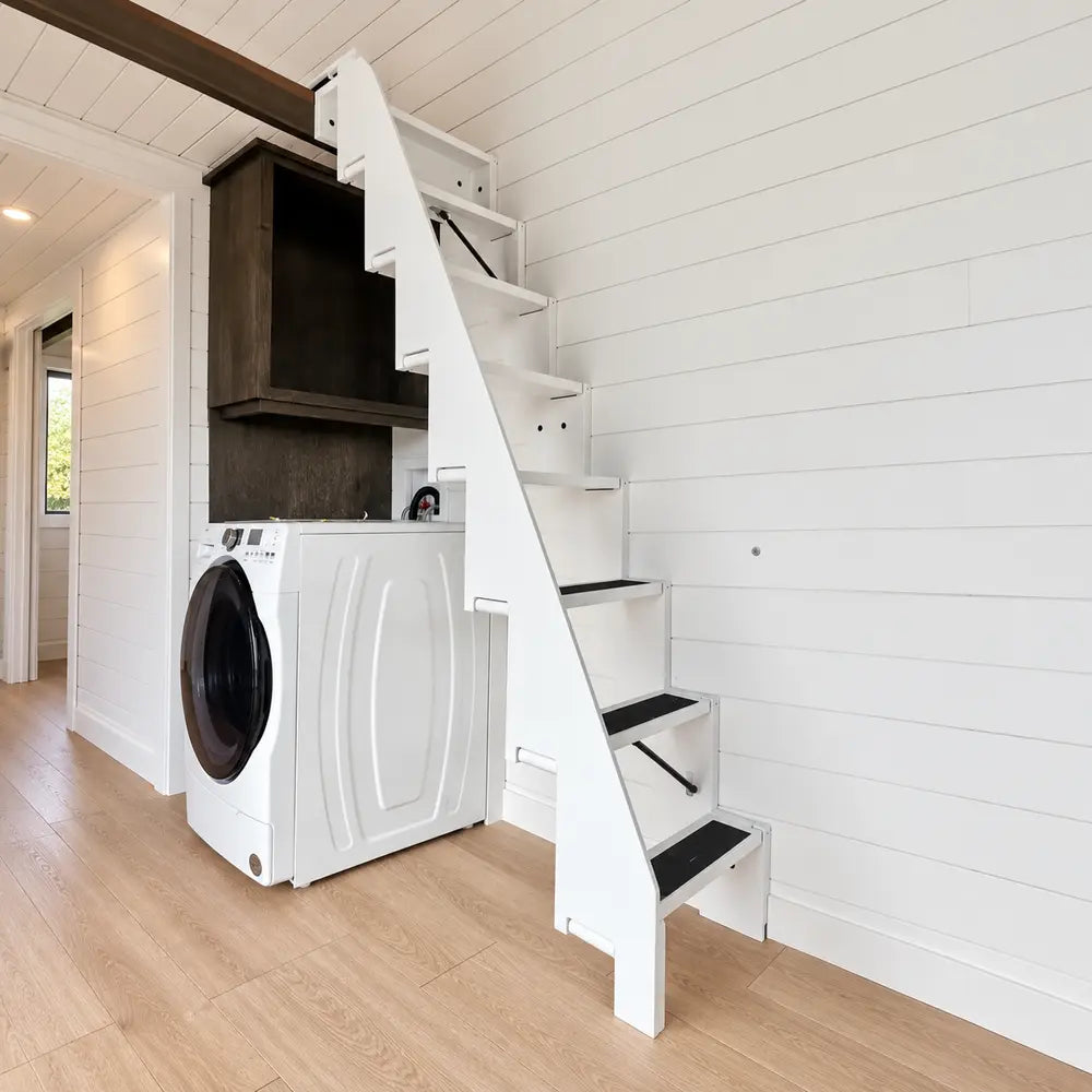 Compact white staircase with dark step grips rising above a washer-dryer inside the bright Wanderlove Tiny Home.