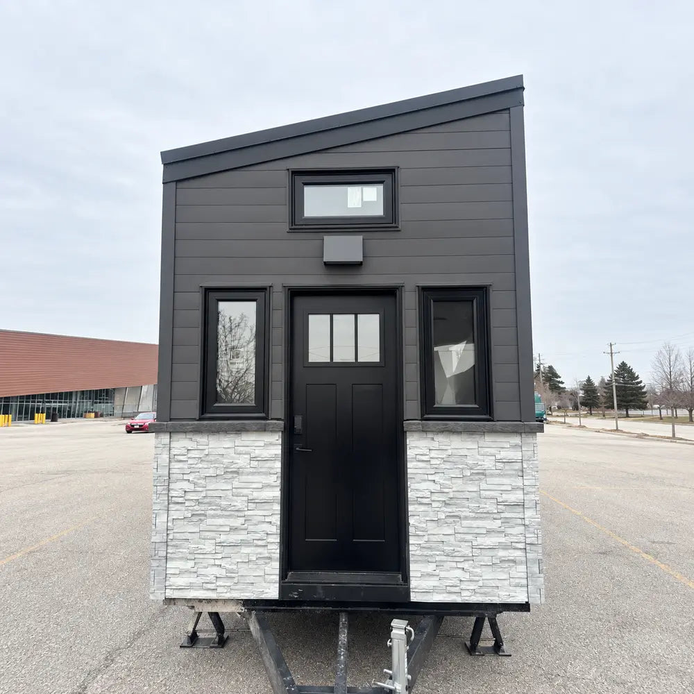 Head-on view of the Wanderlove Tiny Home with a central black door, matching side windows, dark siding, and a light faux-stone lower façade.