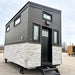 Front view of the Wanderlove Tiny Home featuring a tall black entry door, narrow side windows, transom window, stone siding, and entry steps.