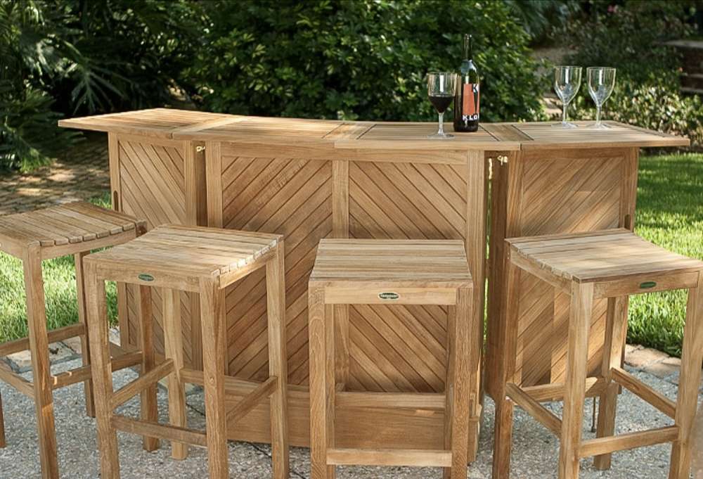 Westminster Teak 5pc Somerset Teak Bar & Stool Set outdoor setup with four stools and a bar topped with wine glasses and a bottle, set against lush tropical plants