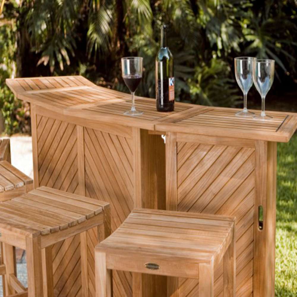 Westminster Teak 5pc Somerset Teak Bar & Stool Set placed on a patio surrounded by greenery with stools tucked under the bar and drinks on top