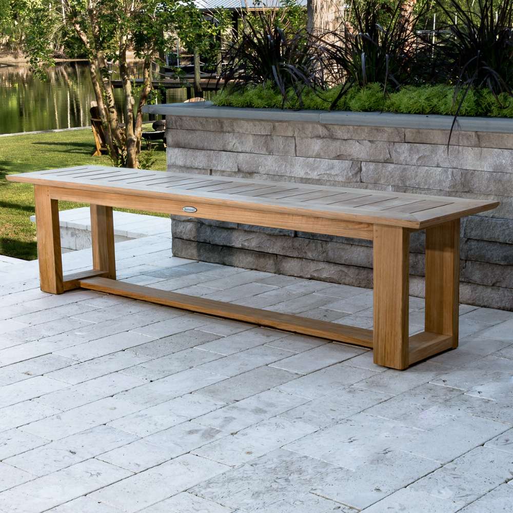 Westminster Teak 6ft Horizon Teak Backless Bench is placed on a stone patio beside a raised planter, with a peaceful lakeside garden and trees behind it