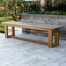 Westminster Teak 6ft Horizon Teak Backless Bench is placed on a stone patio beside a raised planter, with a peaceful lakeside garden and trees behind it