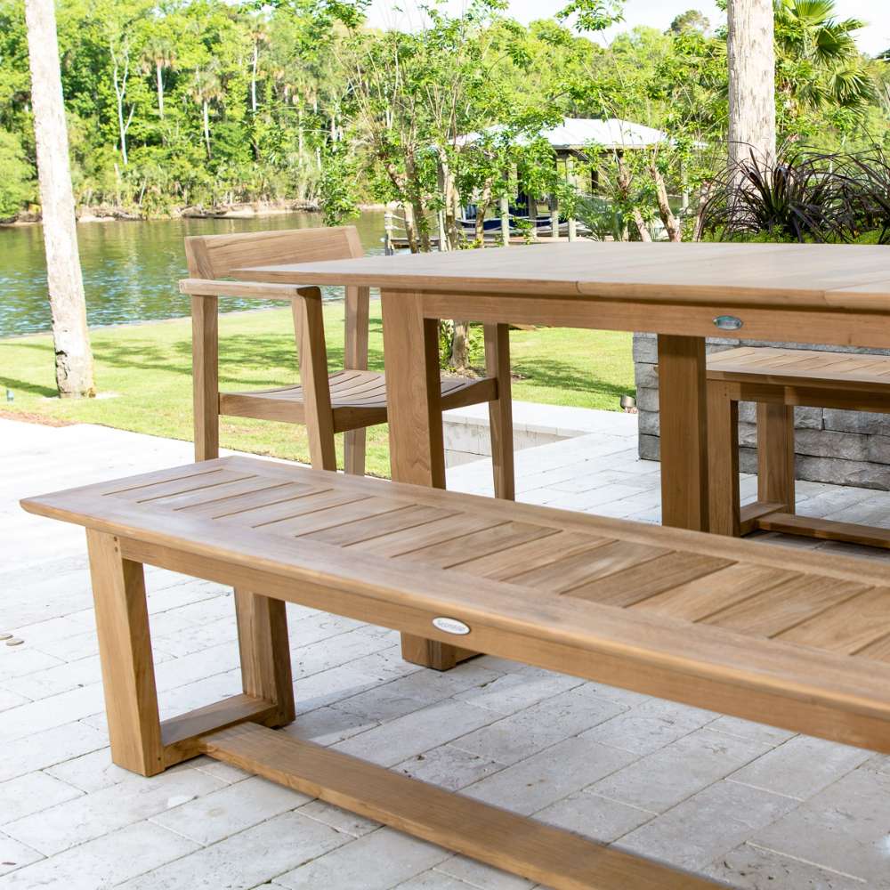 Westminster Teak 6ft Horizon Teak Backless Bench is placed outdoors beside a matching table near a calm lake, surrounded by green trees and grass