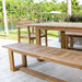 Westminster Teak 6ft Horizon Teak Backless Bench is placed outdoors beside a matching table near a calm lake, surrounded by green trees and grass