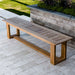 Westminster Teak 6ft Horizon Teak Backless Bench sits alone on a stone patio, surrounded by a landscaped garden with grass and plants