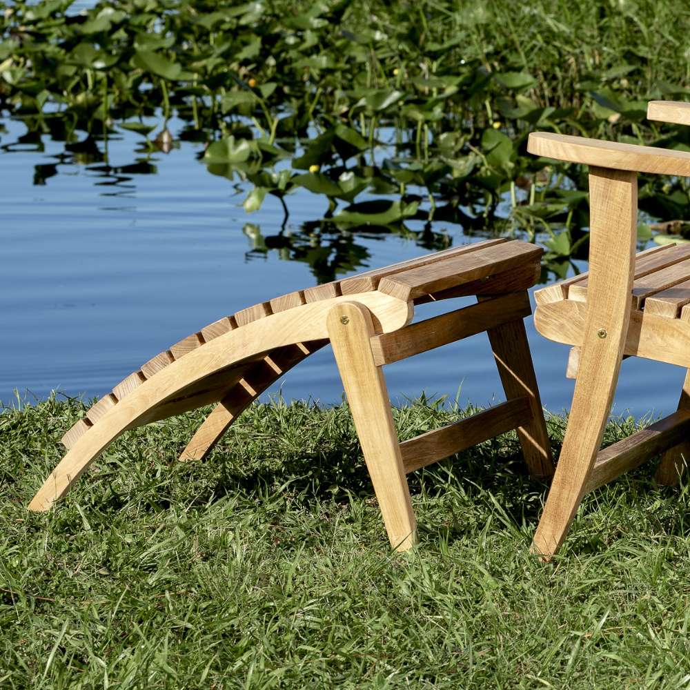 Westminster Teak Adirondack Footrest placed outdoors beside an Adirondack chair near water, showing a relaxing outdoor setup