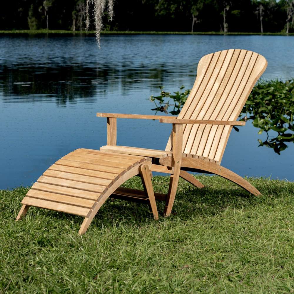 Westminster Teak Adirondack Lounger and Ottoman sits on grass by a lake, featuring a curved teak recliner with a matching slatted footrest